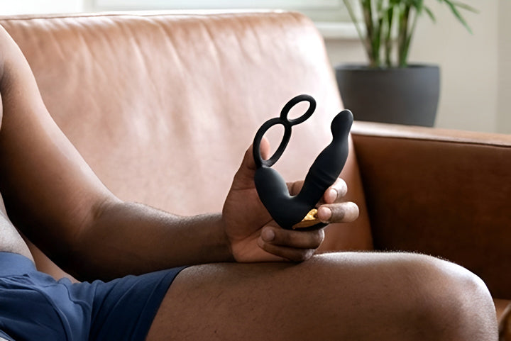 A man relaxing on a leather sofa at home, holding the black silicone Prostate Massager with Dual Performance Rings.